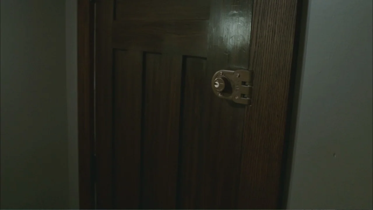 The Stepfather (2009) movie still: closeup — A close view of a dark wood-paneled door mounted with a Yale deadbolt lock. The door is set within a…