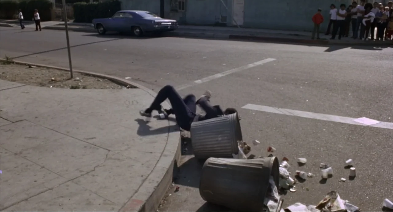 Foxy Brown (1974) movie still: wide — A suburban street intersection features a concrete sidewalk, an asphalt road, and a parked car. A person falls…