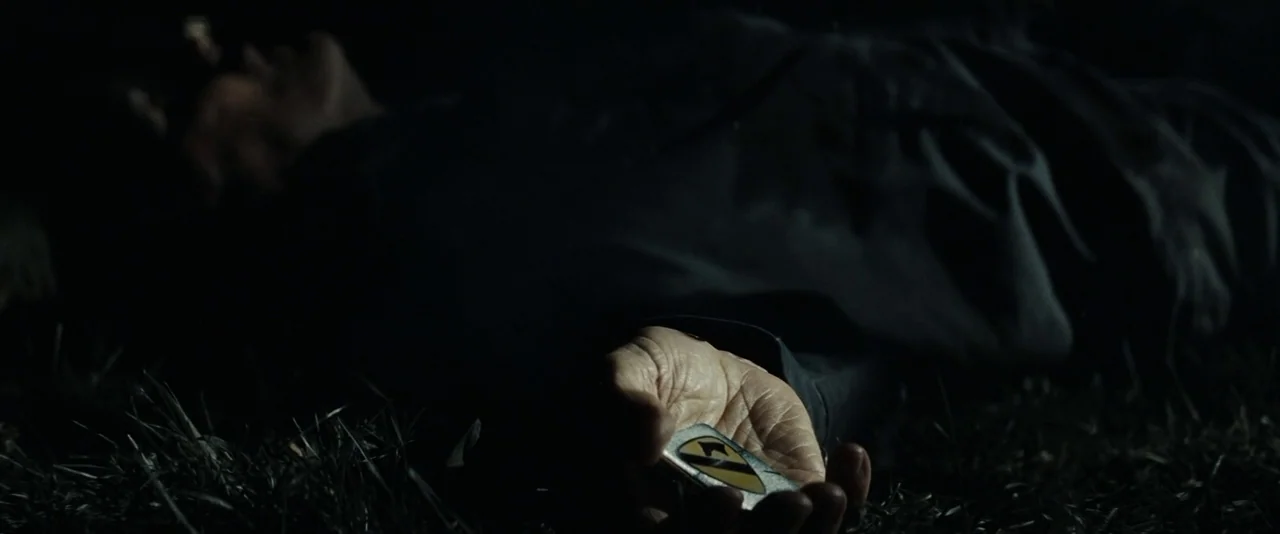 Gran Torino (2008) movie still: closeup — A person lies prone on the dark ground covered in blades of grass. The individual holds a metallic patch…