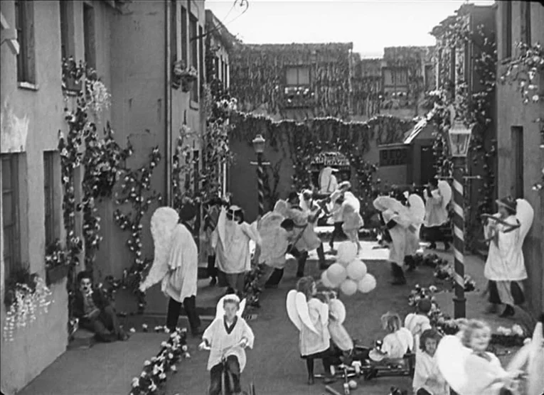 The Kid (1921) movie still: wide — A narrow street set is decorated with vines and flowers on the building facades. Numerous people wearing white…