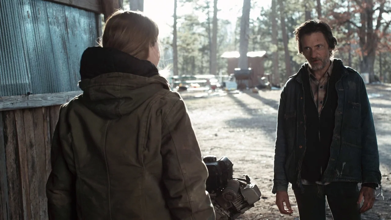 Winter’s Bone (2010) movie still: medium — A clearing surrounded by pine trees with a rustic wooden shed structure to left. A woman in a green coat…