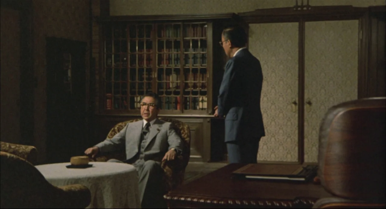 The Return of Godzilla (1984) movie still: medium — A dimly lit room features dark wood bookshelves and patterned wallpaper on the walls. One man…