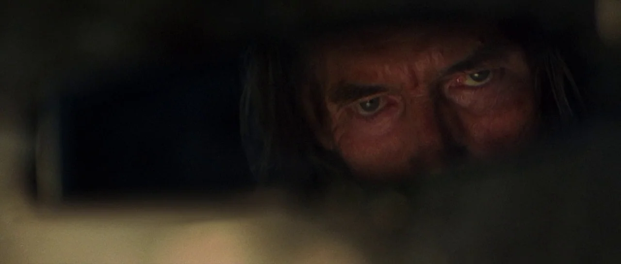 The Hills Have Eyes (2006) movie still: closeup — An older man gazes intently from the darkness through a narrow, horizontal aperture. Only his…