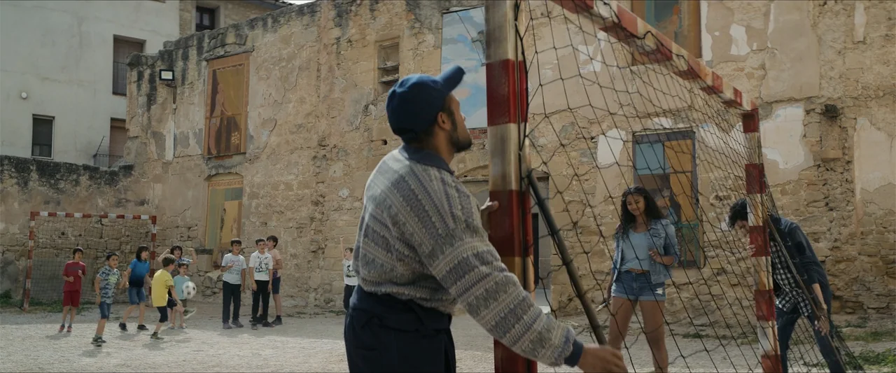 A Hipster in Rural Spain (2024) movie still: medium — An outdoor dirt area features a weathered stone wall with painted murals behind a goal. A man…