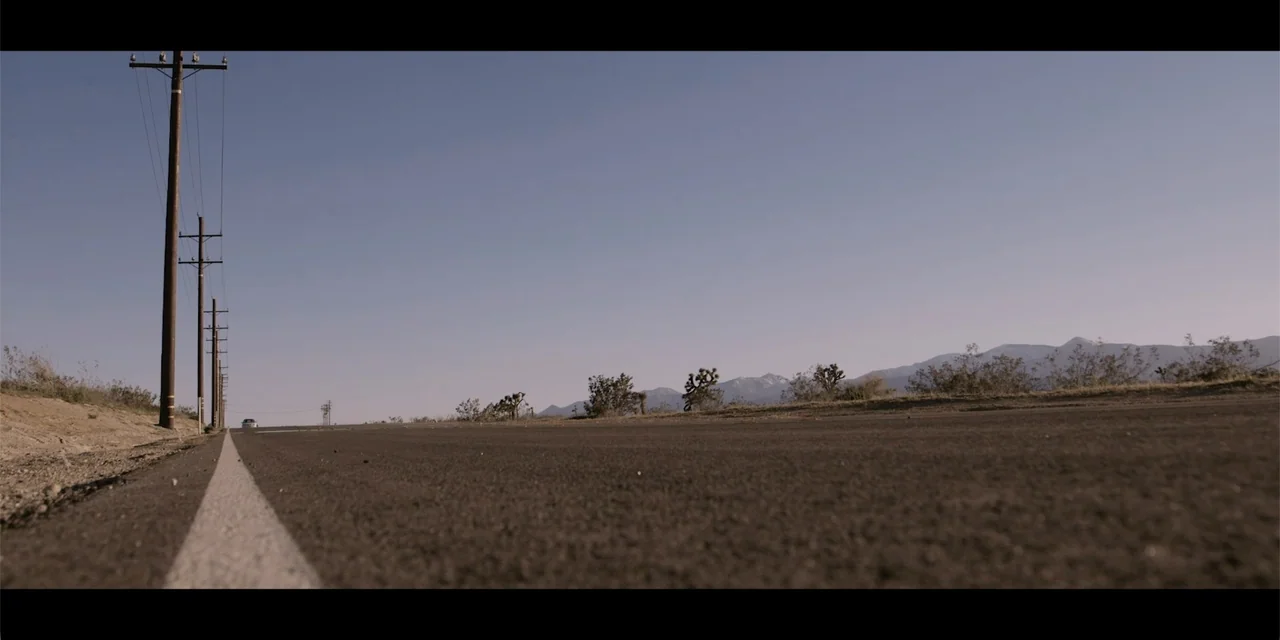 La coda del diavolo (2024) movie still: establishing — A flat asphalt desert road stretches into the distance under a clear blue sky. Telephone…