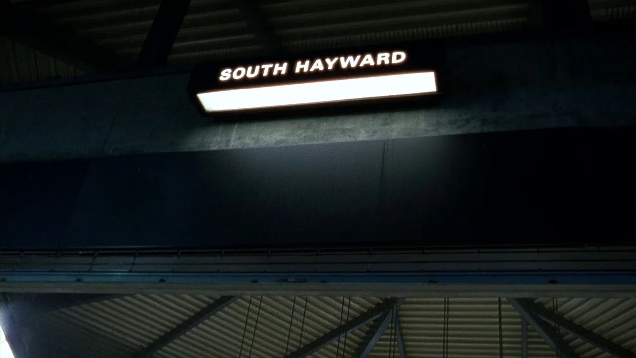 Fruitvale Station (2013) movie still: establishing — The illuminated station sign for South Hayward is mounted on a dark concrete structure. A…