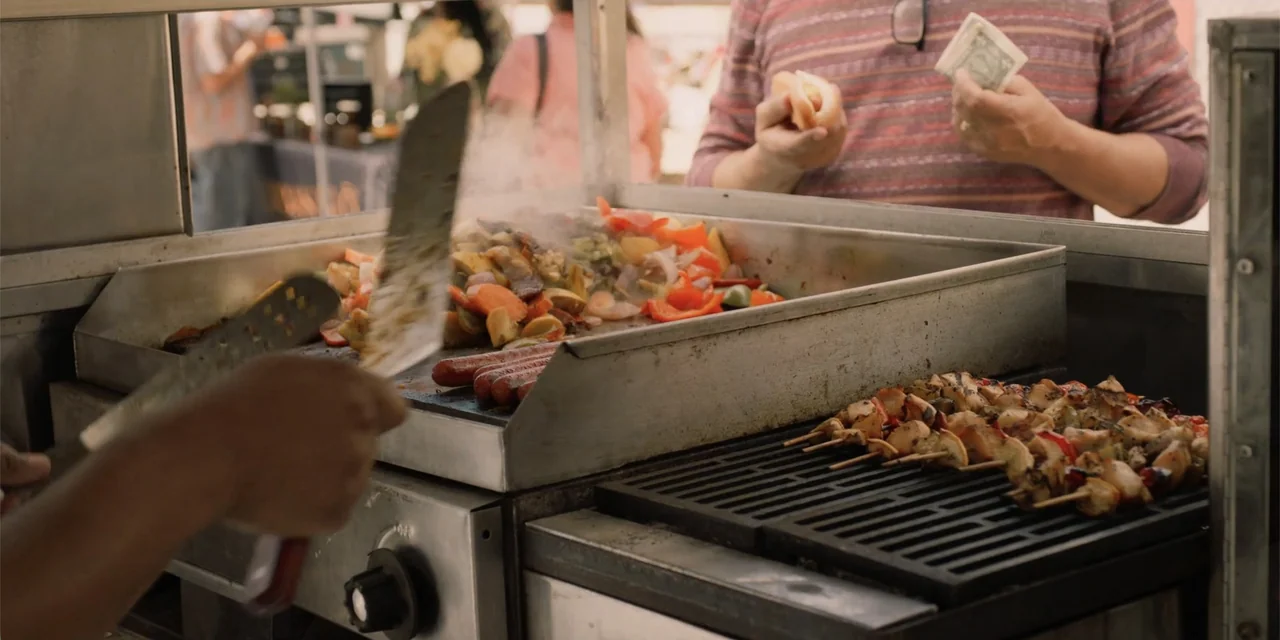 Música (2024) movie still: medium — A stainless steel food cart holds a hot griddle filled with chopped vegetables and sausages. A customer standing…