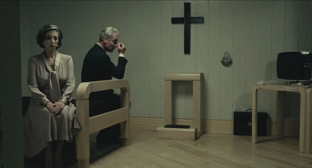 Todo Modo (1976) movie still: medium — A spartan room features a wooden bench, a kneeling stool, and a wall-mounted cross. A woman and a man sit…