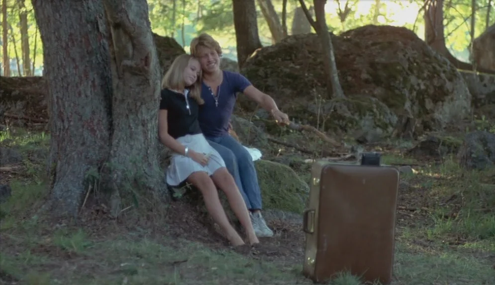 A Swedish Love Story (1970) movie still: fullbody — A teenage boy and girl rest against a large tree in a wooded area. A brown vintage suitcase sits…