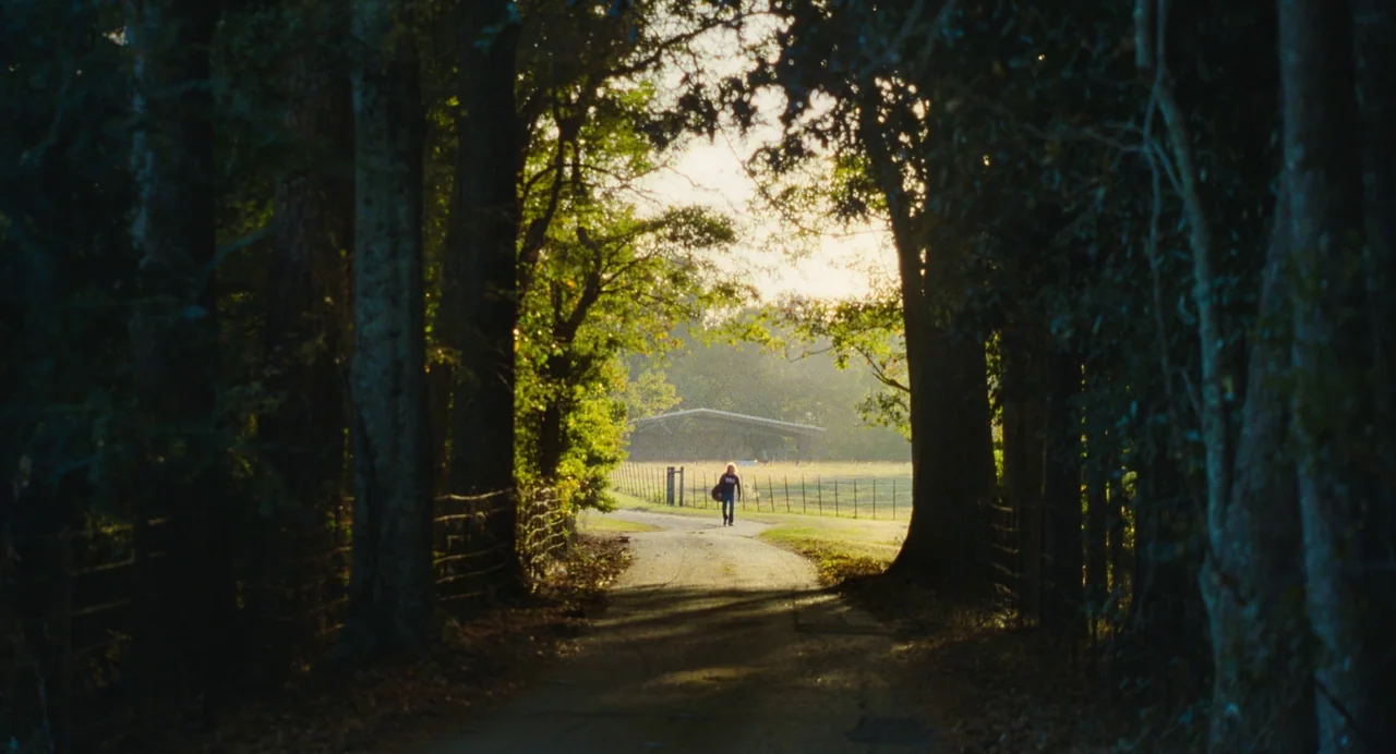 The Iron Claw (2023) movie still: wide — A narrow dirt road flanked by tall trees leads toward an open fenced pasture. A lone figure walks away from…