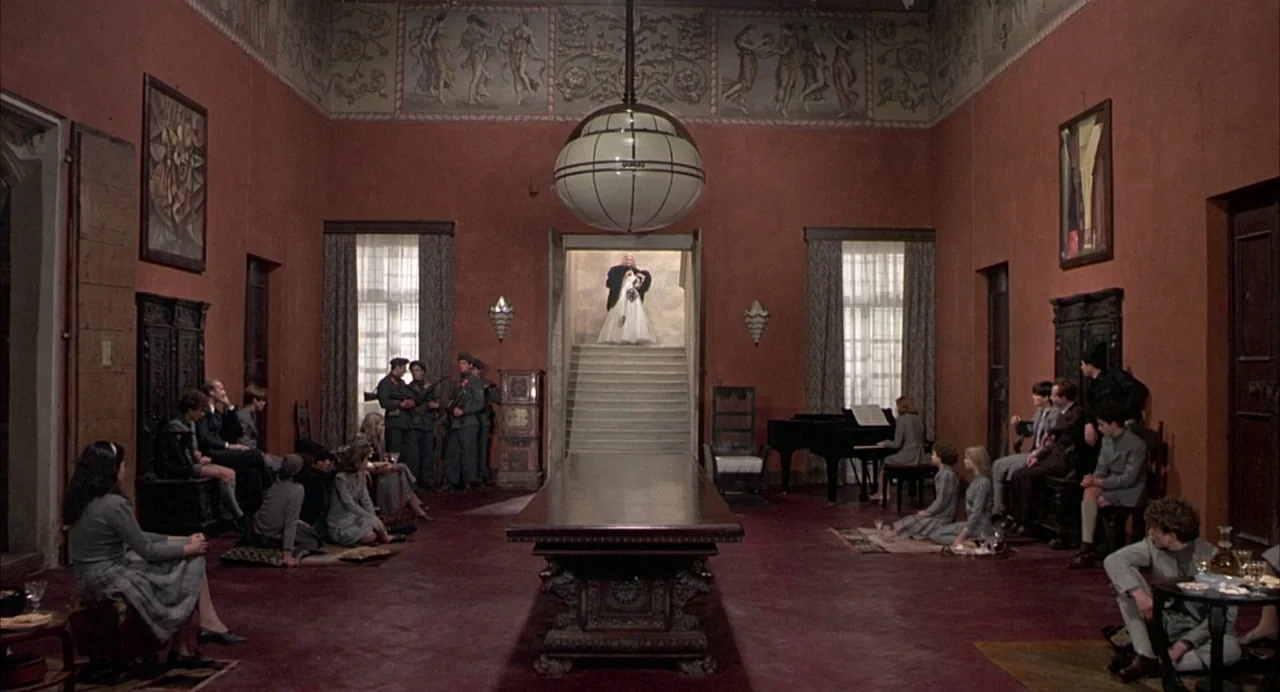 Salò, or the 120 Days of Sodom (1975) movie still: wide — A spacious room with terracotta-colored walls and a decorative frieze along the ceiling.…