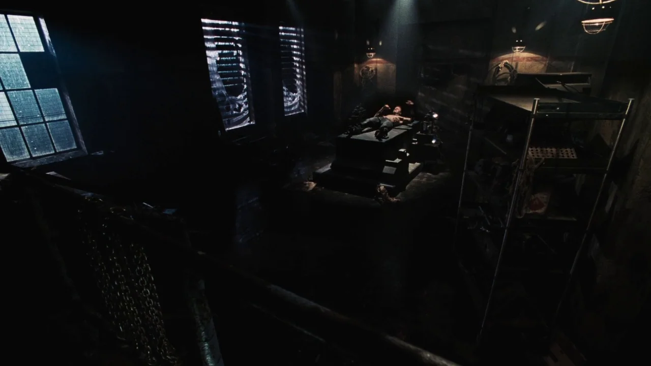 Saw V (2008) movie still: wide — A dimly lit, industrial-style room features metal surfaces, chains, and grated windows. A man lies on a flat metal…