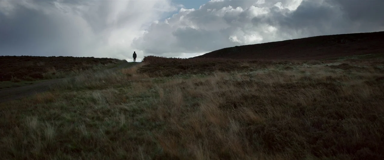 Ghost Stories (2017) movie still: wide — An expansive, windswept moorland under a heavy, overcast sky with rolling hills. A lone figure stands on…