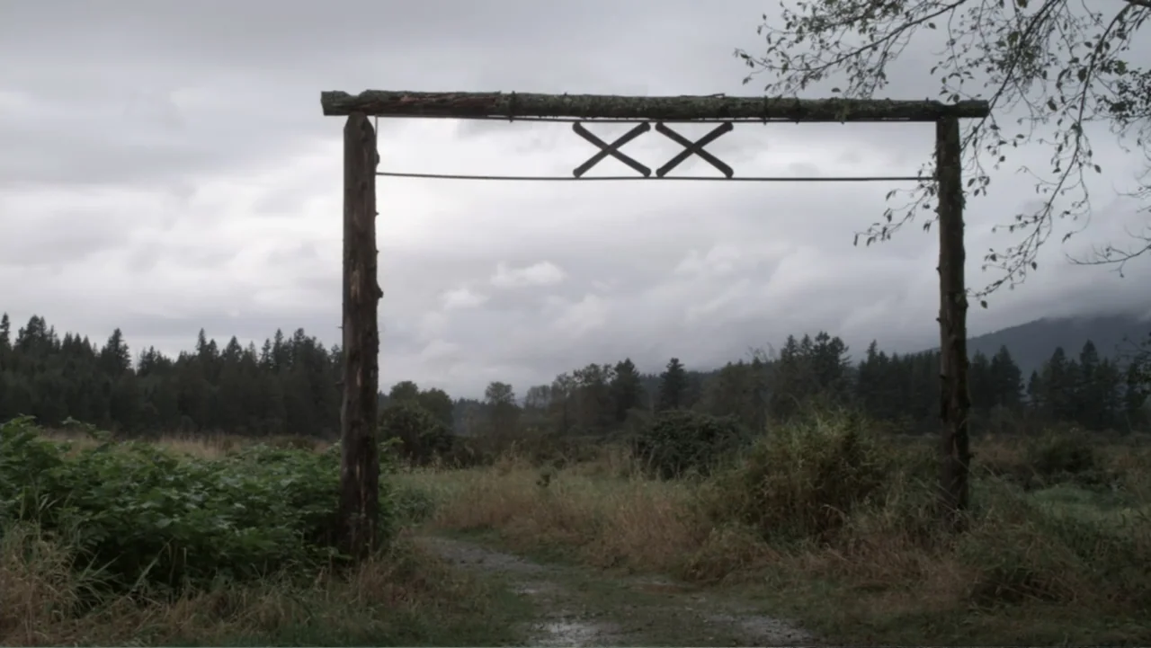 Dawn Rider (2012) movie still: establishing — A rural wooden gate with crossed beams marks the entrance to a damp grassy field. A worn dirt path…