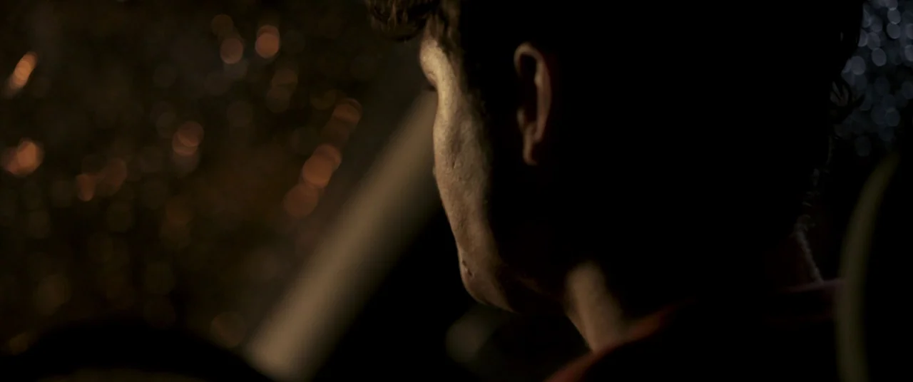 Talk To Me (2022) movie still: closeup — The interior of a vehicle at night with blurred street lights visible outside. A man in his twenties sits…