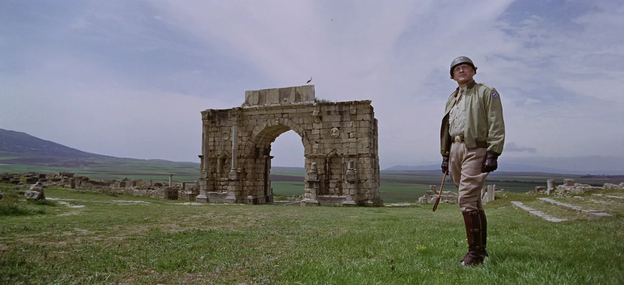 Patton (1970) movie still: fullbody — A grassy field contains the stone remnants of an ancient Roman archway. A uniformed officer stands in the…