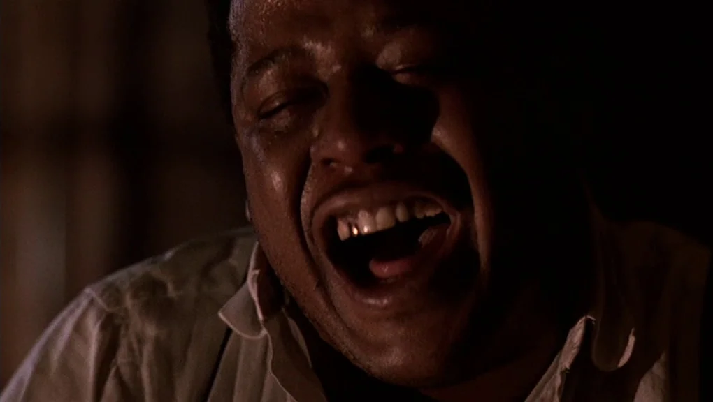 Bird (1988) movie still: closeup — A dark, dimly lit indoor space with undefined surroundings and deep shadows. A man with a gold tooth in his mouth…