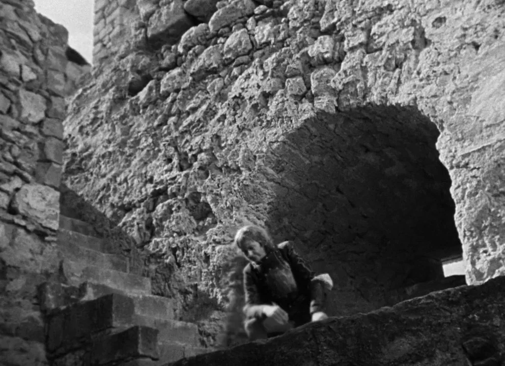 Othello (1952) movie still: wide — Ancient stone ruins featuring rough-hewn masonry and a weathered stone staircase. A lone figure crouches on a…