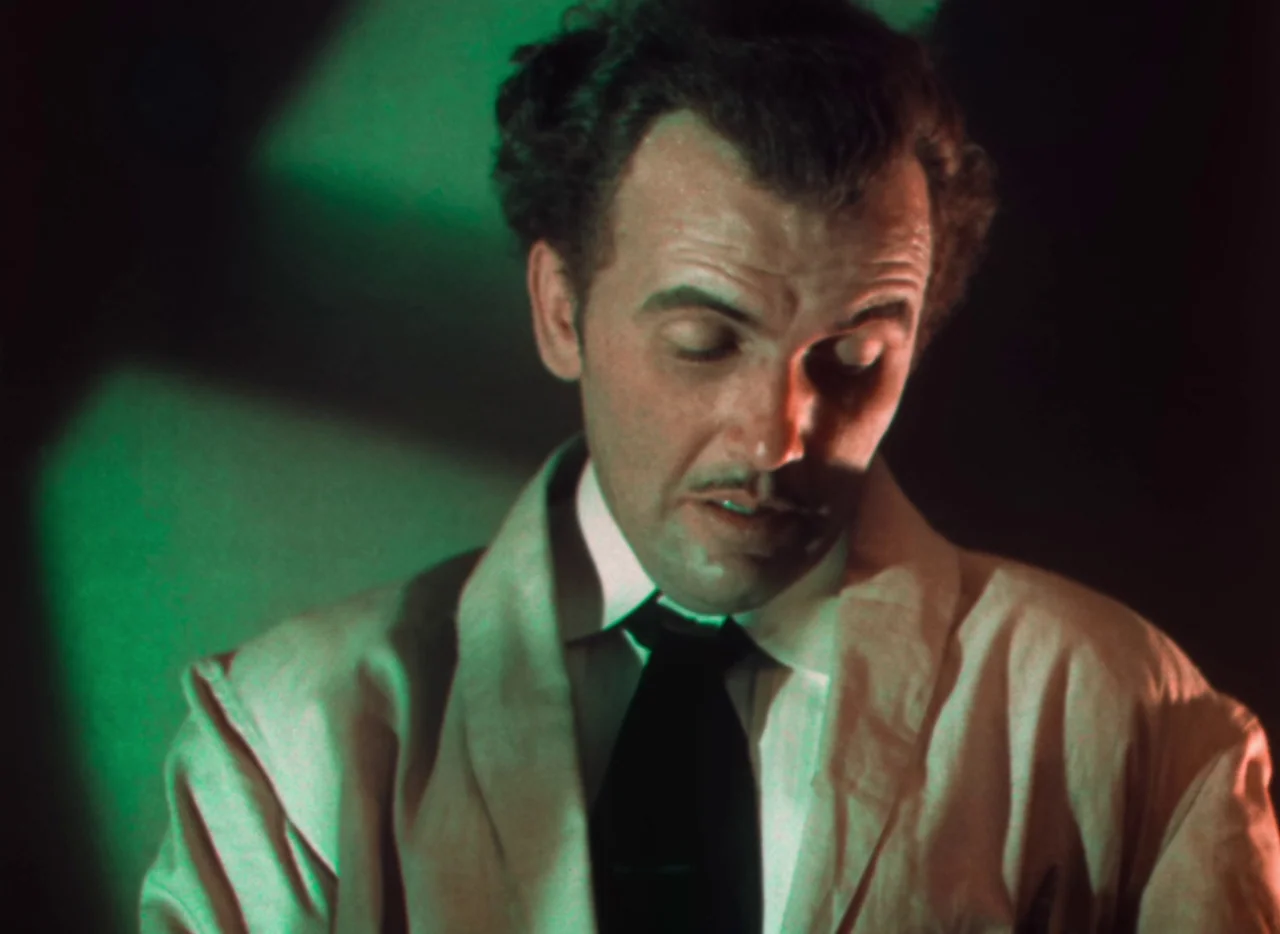Doctor X (1932) movie still: closeup — A dimly lit laboratory interior is illuminated by a sharp green light source. A man in a lab coat looks down…