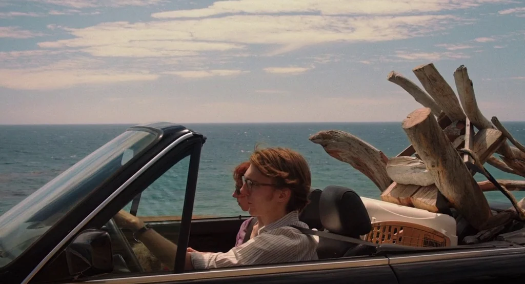 Ruby Sparks (2012) movie still: medium — A convertible car travels along a road next to the bright blue ocean. The backseat of the vehicle is loaded…