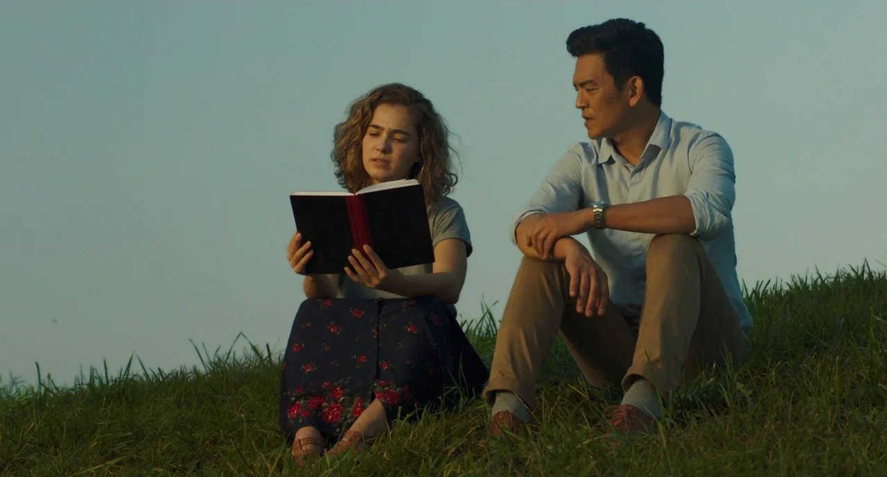 Columbus (2017) movie still: medium — A man and a woman sit together in the grass at golden hour. The woman reads from a black book while the man…