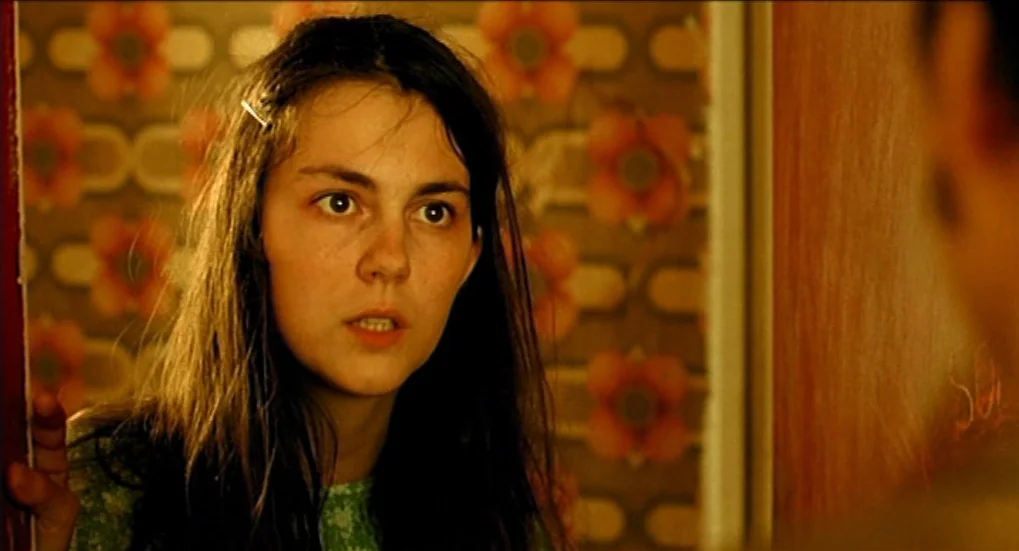 Jeux d’Enfants (2003) movie still: closeup — A dimly lit interior space features retro floral-patterned wallpaper covering the walls. A young woman…