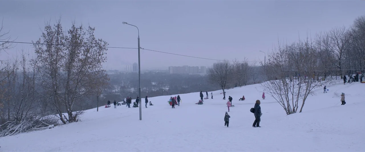 Loveless (2017) movie still: wide — A snow-covered public park hill overlooks a distant city skyline under overcast skies. Small groups of people…