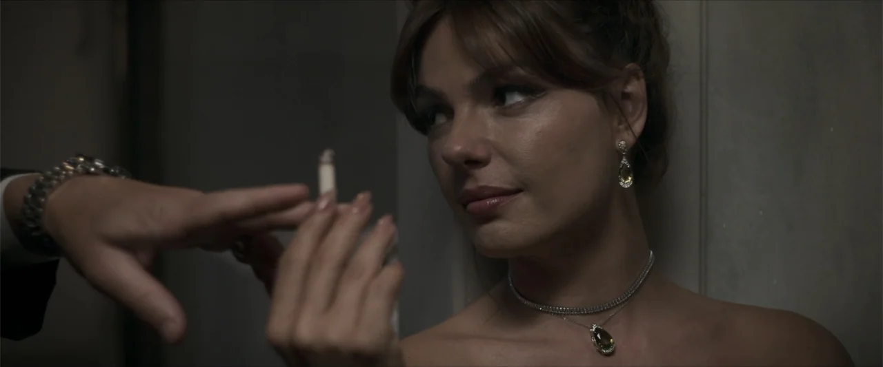 Angela (2023) movie still: closeup — A dimly lit interior space with plain walls and visible shadows. A woman in formal jewelry looks toward a hand…