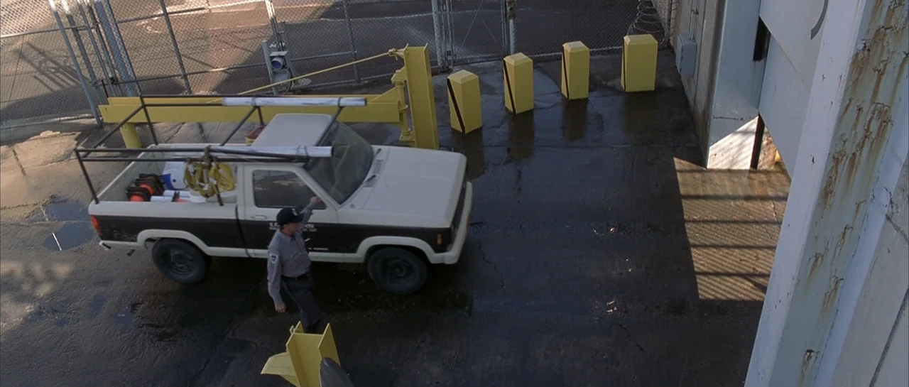 Bandits (2001) movie still: wide — A concrete utility lot features a white and brown pickup truck near a fence. A uniformed guard stands beside the…