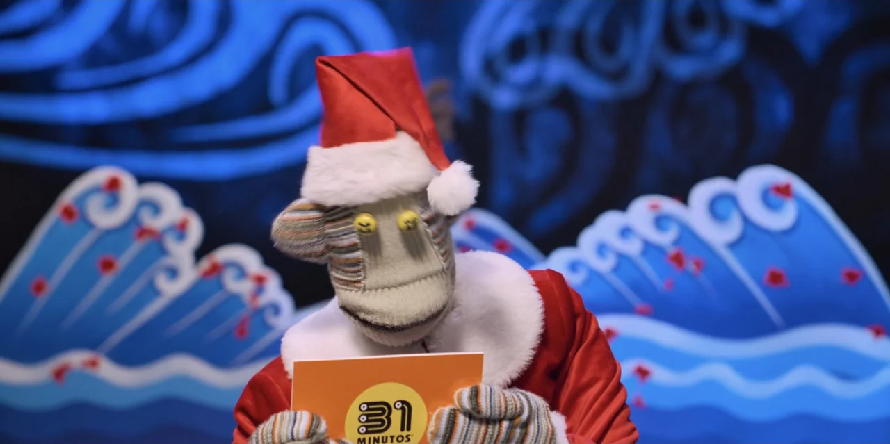 31 Minutes: One Hot Christmas (2025) movie still: closeup — A puppet sits in a studio with stylized blue wave cutouts in the background. The puppet…