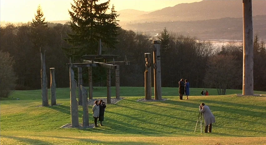 Double Happiness (1994) movie still: wide — A spacious green park features several tall carved wooden poles and a geometric structure. People walk…