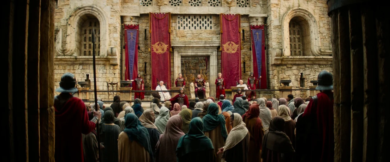 The Book of Clarence (2023) movie still: wide — An ancient stone colosseum features a raised platform decorated with large vertical crimson banners.…