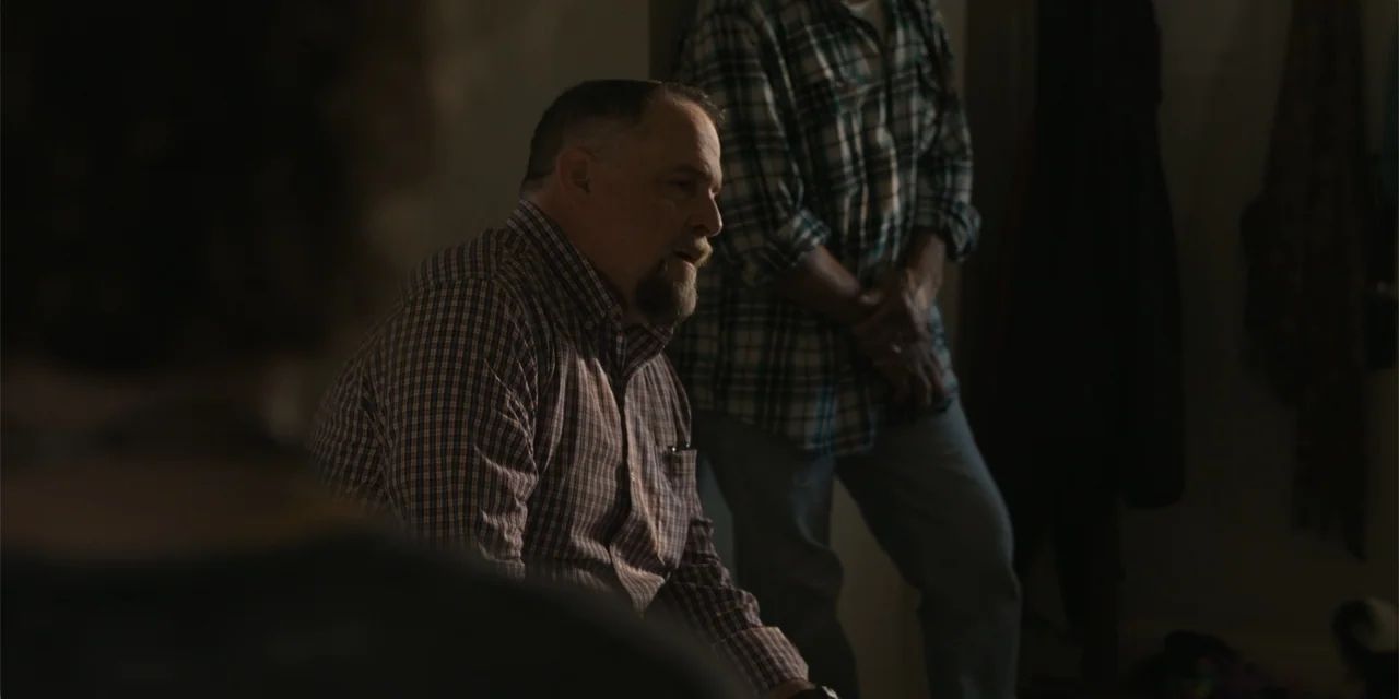 Caddo Lake (2024) movie still: medium — A dimly lit interior room with neutral walls and dark shadow areas. One man sits in the foreground speaking…