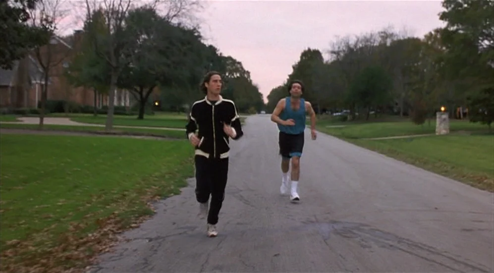 Bottle Rocket (1996) movie still: fullbody — A quiet residential street lined with green lawns and large trees at dusk. Two young men jog…