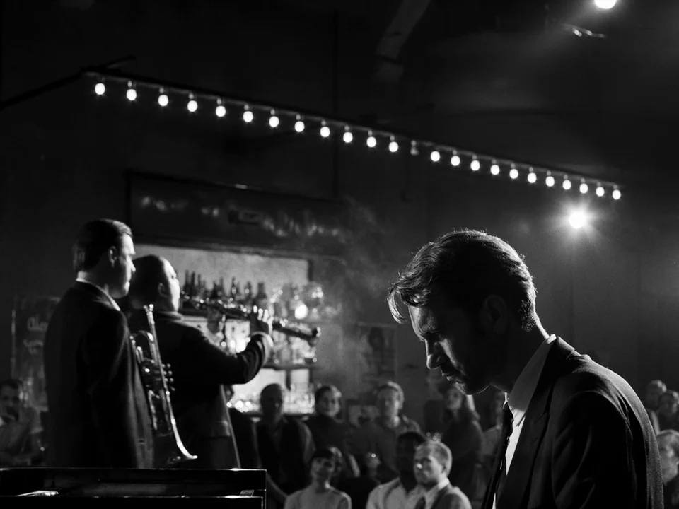 Cold War (2018) movie still: medium — A dimly lit jazz club with a row of illuminated bulbs hanging above. A musician sits at a piano in the…