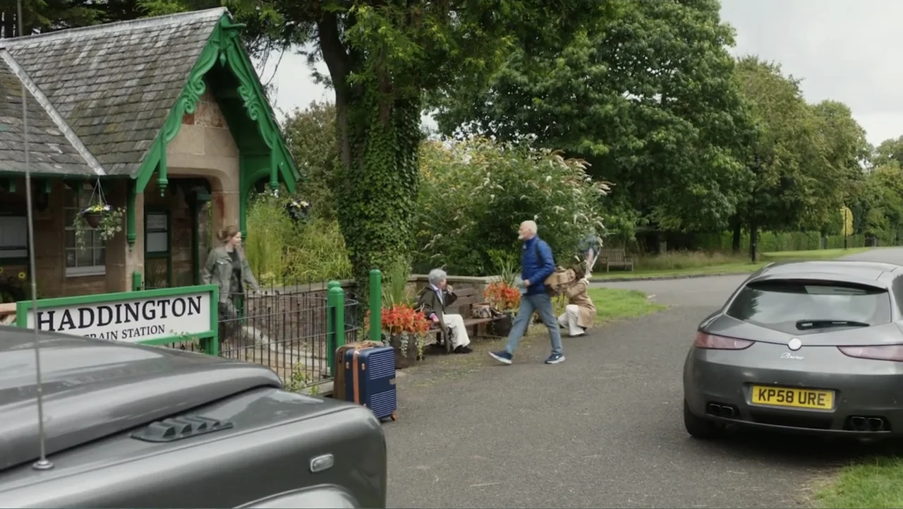 A Scottish Love Scheme (2024) movie still: wide — A stone train station building sits amidst green foliage and trees in the countryside. A group of…