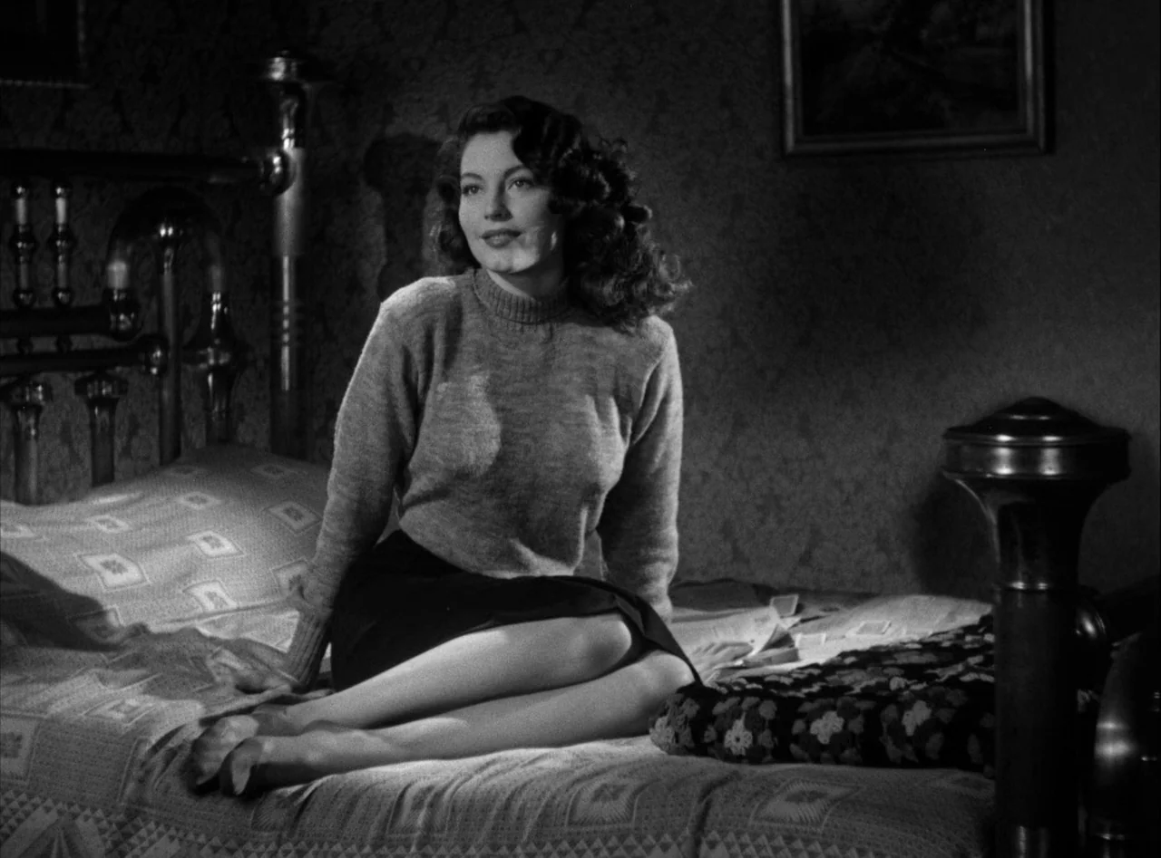 The Killers (1946) movie still: medium — A dark, textured bedroom interior with ornate metal bed frame and patterned bedding. A woman in a dark…