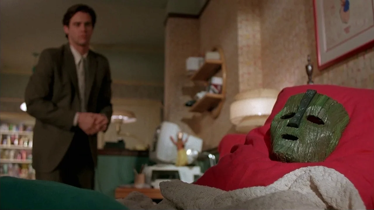 The Mask (1994) movie still: medium — A man stands in the background of a furnished apartment room. A carved wooden green mask rests in the…