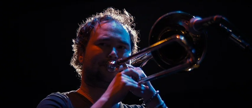 This Movie Is Broken (2010) movie still: closeup — A man performs on a dark stage with minimal ambient background. He holds a brass trombone to his…