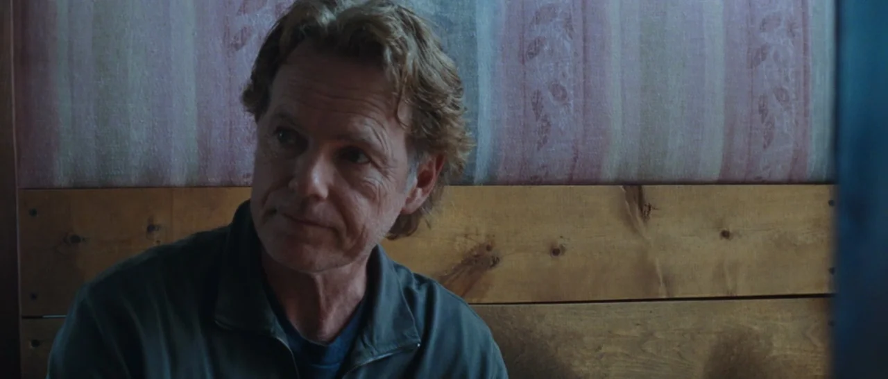 Wildlike (2014) movie still: medium — A man sits in a booth with a wooden backrest and patterned wallpaper behind him. He is looking off to the side…