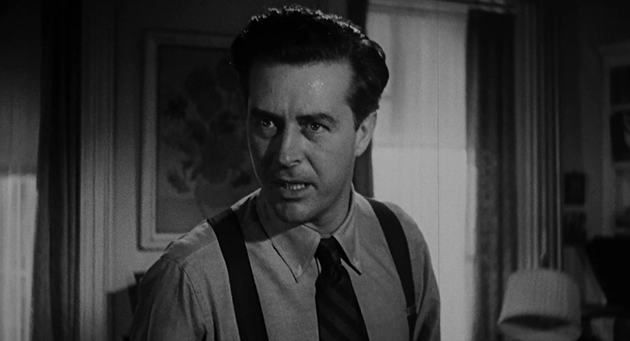 Dead Men Don’t Wear Plaid (1982) movie still: medium — A man stands in an interior room with a framed floral print on the wall. He wears a dress…