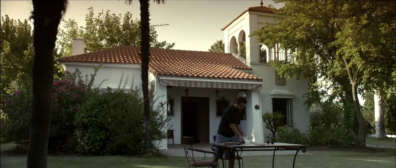 Otro verano (2013) movie still: wide — A white stucco house with a red tile roof sits behind a grass lawn. A man stands at a table on the patio…