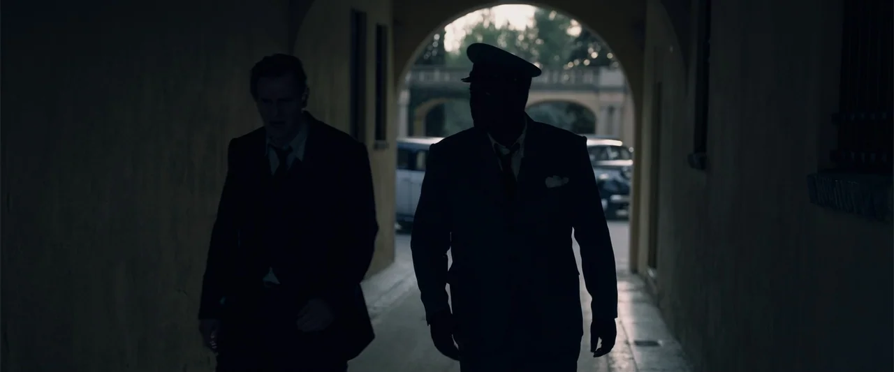 Marlowe (2023) movie still: fullbody — A stone arched walkway features a shadowed corridor and a bright sunlit opening beyond. Two men walk side by…