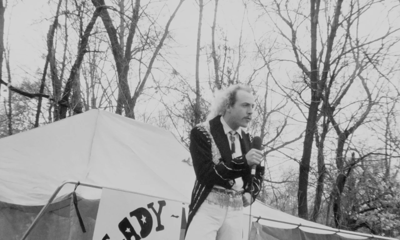Multiple Maniacs (1970) movie still: medium — An outdoor fairground setting features a white tent positioned before a line of bare trees. A man in a…