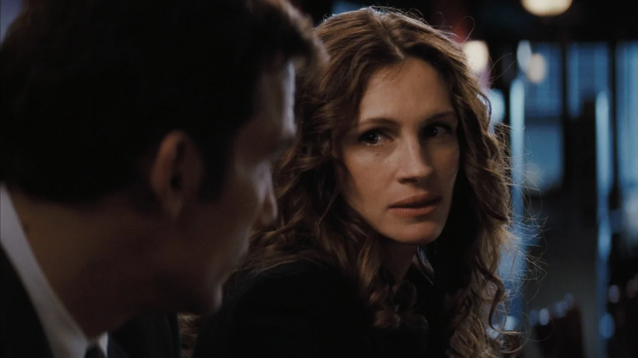 Duplicity (2009) movie still: medium — A dimly lit indoor space with blurred background lights visible behind the subjects. A woman with long brown…