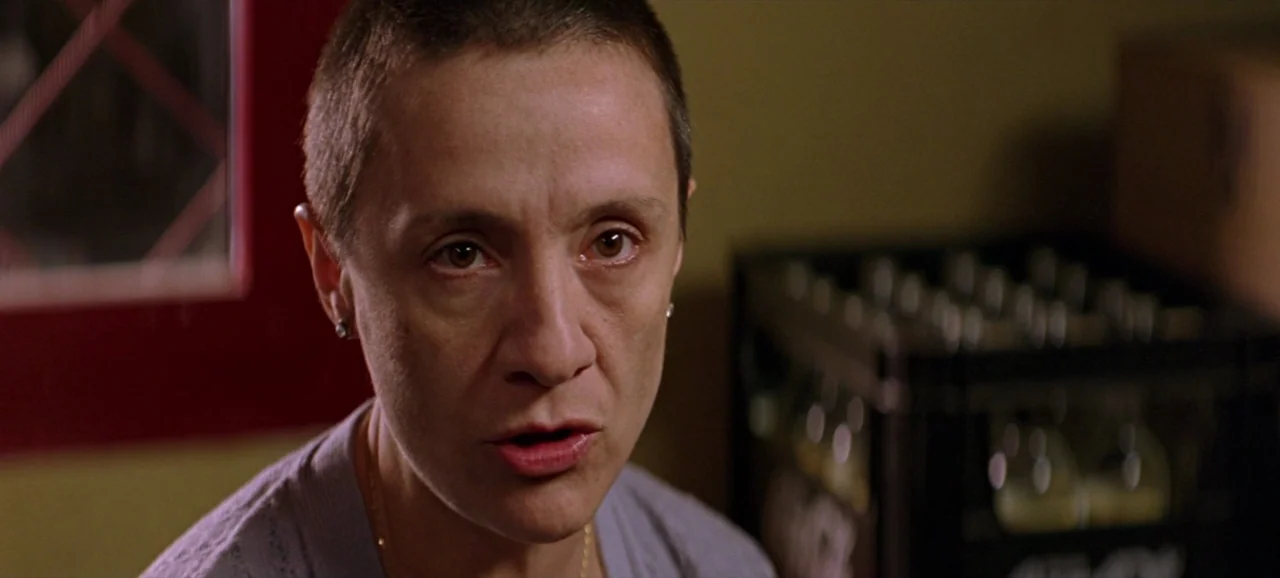 Volver (2006) movie still: closeup — A dark, dimly lit store room contains a plastic crate of glass bottles. A woman with short cropped hair speaks…
