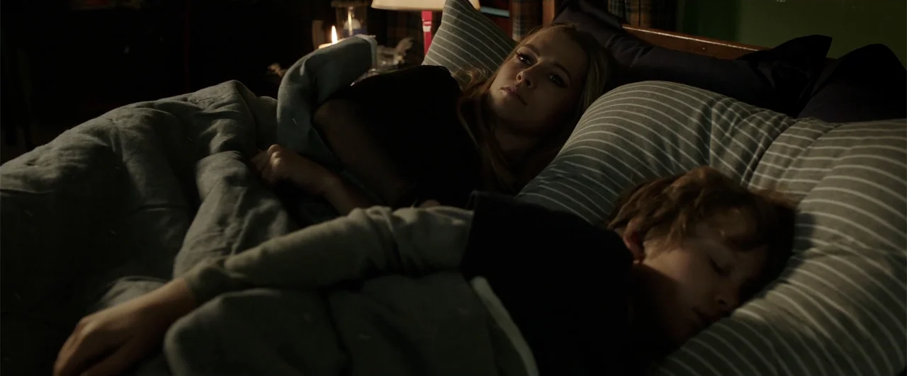 Lights Out (2016) movie still: medium — A dimly lit room contains a bed with gray striped pillows and blankets. A woman lies watching a sleeping…