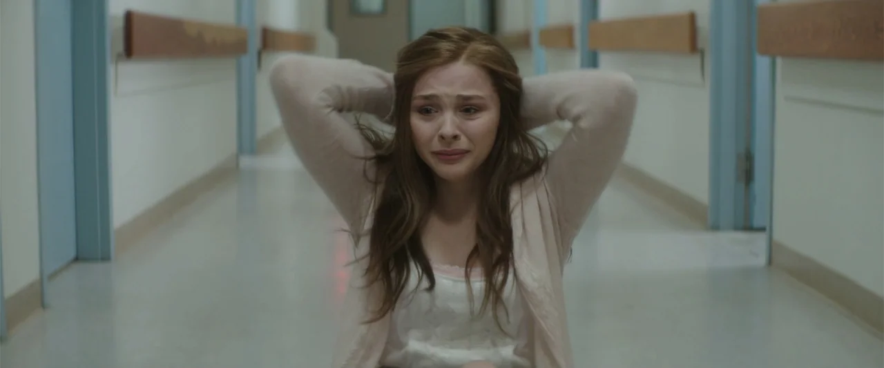 If I Stay (2014) movie still: medium — A sterile hospital corridor with pale walls and blue doors stretches into the distance. A young woman kneels…
