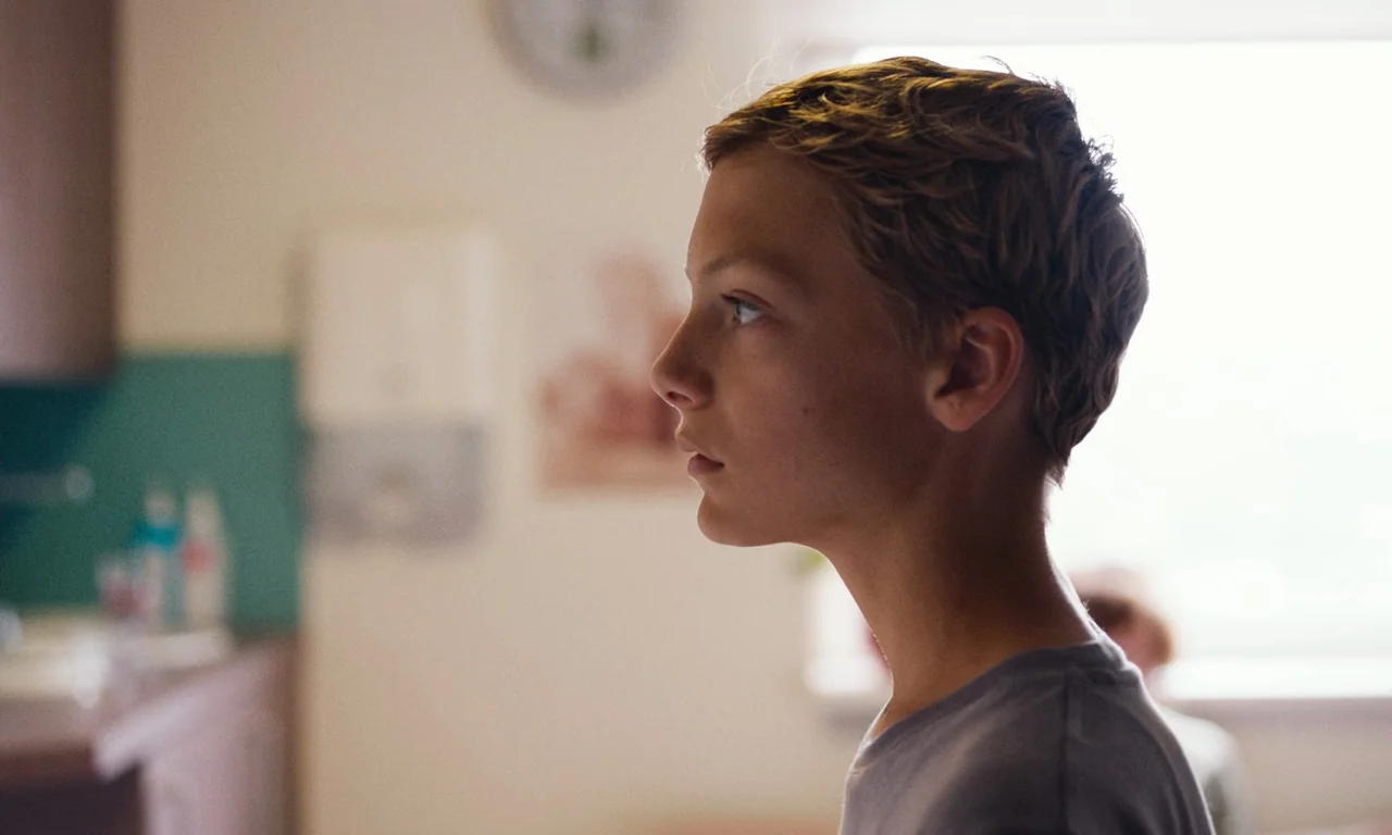 Close (2022) movie still: closeup — A light-colored room with a blurred turquoise backsplash and a wall clock. The boy is in profile looking toward…