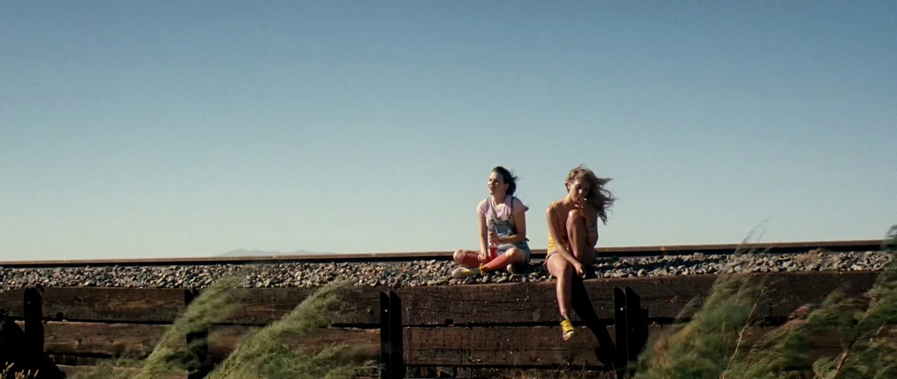 Little Birds (2011) movie still: wide — Two women sit perched on dark wooden railroad ties atop a gravel embankment. They are positioned against a…